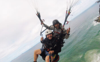 instrutor e cliente sorindo para a foto durante um voo de parapente, com o céu nublado e mar do Rio de Janeiro abaixo