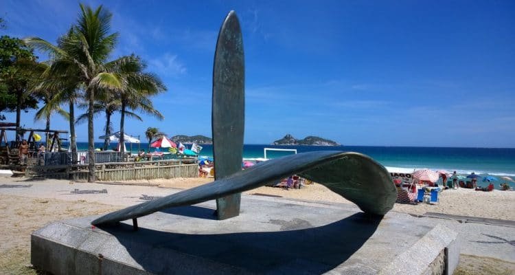 escultura de prancha e asa delta na orla do Rio de Janeiro em homenagem a Pepê Lopes, com palmeiras no fundo à esquerda, pessoas tomando sol mais ao fundo e céu azul e mar