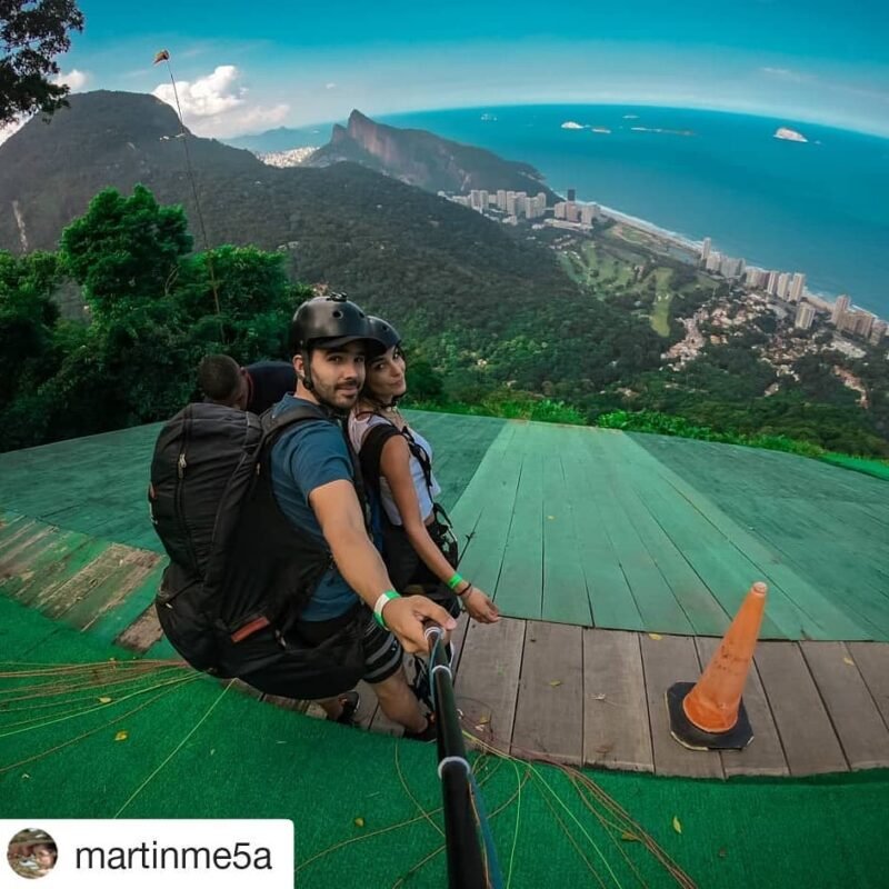 selfie de um homem e uma mulher se preparando para realizar o salto de parapente na Rampa de Vôo Livre da Pedra Bonita, com a cidade do Rio de Janeiro ao fundo, mar e céu azul